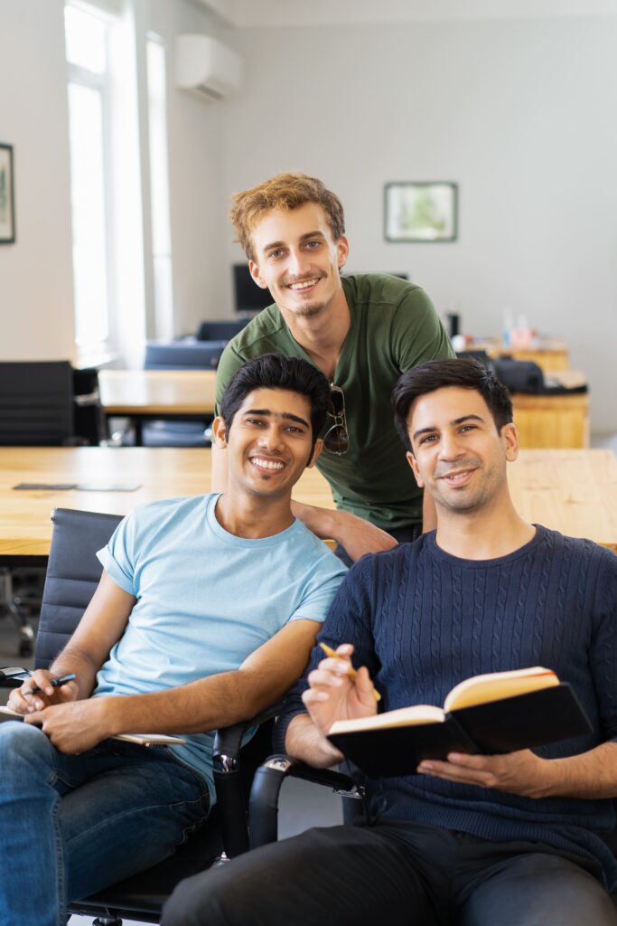 Three contented students sitting side by side with a laptop and books, grinning and focusing on the camera, represent cooperation and a productive study atmosphere following credit transfer.